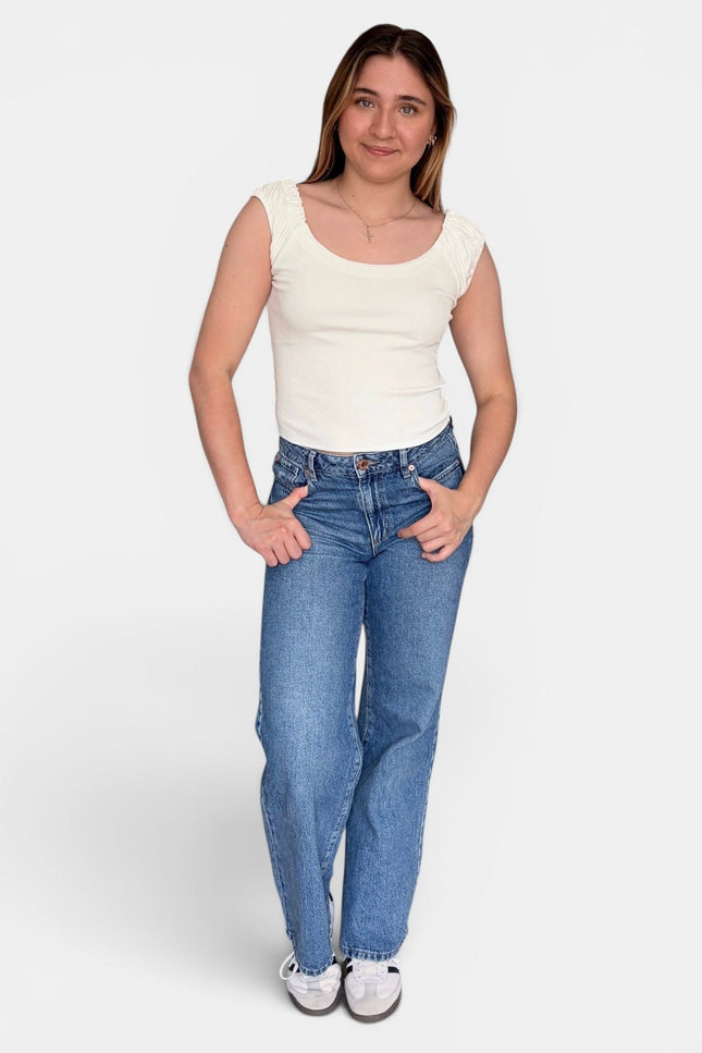A woman modeling a sleeveless cream top and blue jeans against a light gray studio background.