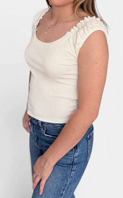 A young woman with blonde hair is wearing a cream-colored sleeveless top with ruffled trim and blue jeans, standing against a plain white studio background.