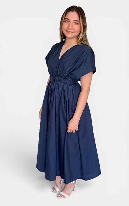 A woman modeling a navy blue midi dress with a V-neckline, short sleeves, and a gathered waist, paired with white heeled sandals against a light gray studio background.