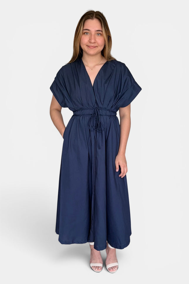 A woman modeling a navy blue maxi dress with short flutter sleeves, a V-neckline, and a self-tie waist belt, set against a plain light gray background.