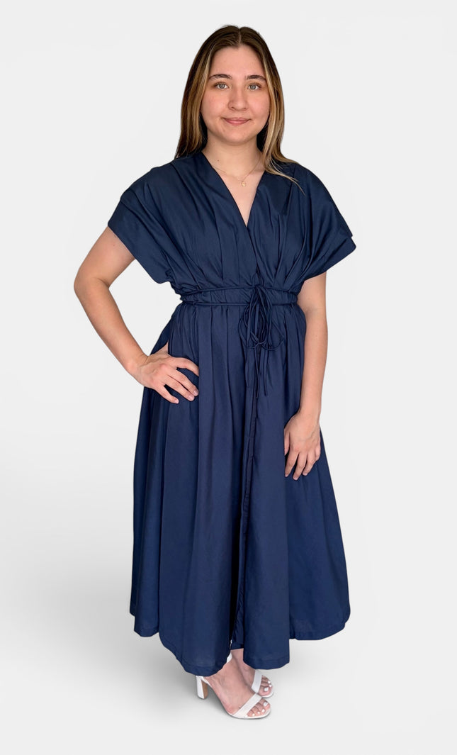 A woman modeling a navy blue midi dress with short flutter sleeves, a V-neckline, and a self-tie waist belt, paired with white heeled sandals against a light gray studio background.