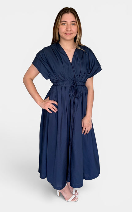 A woman modeling a navy blue midi dress with short flutter sleeves, a V-neckline, and a self-tie waist belt, paired with white heeled sandals against a light gray studio background.
