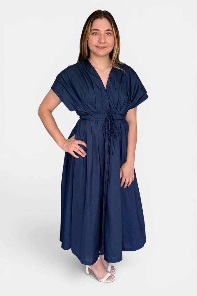 A woman modeling a navy blue midi dress with short flutter sleeves, a V-neckline, and a self-tie waist belt, paired with white heeled sandals against a light gray studio background.