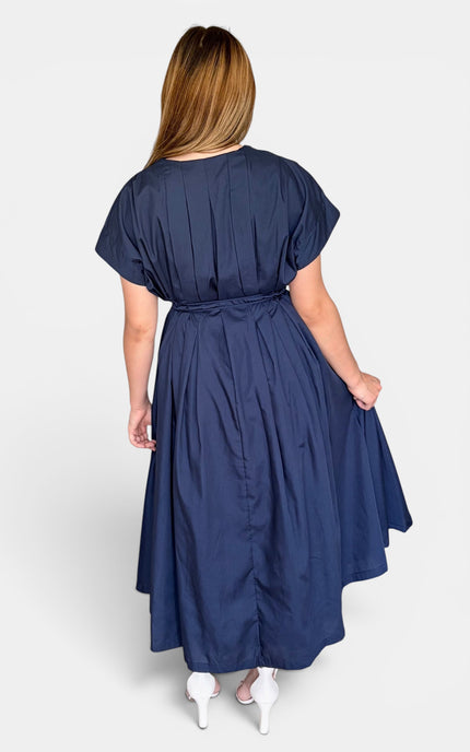 A woman viewed from the back wearing a navy blue midi dress with short sleeves, a gathered waist, and a flared skirt, paired with white heels against a light gray studio background.