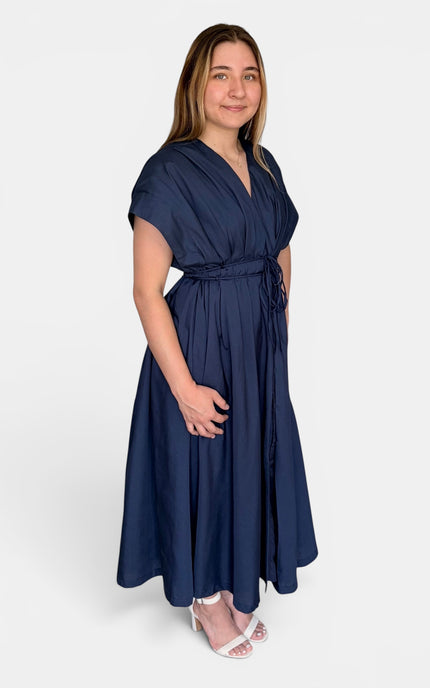 A woman modeling a navy blue maxi dress with short sleeves, a V-neckline, and a matching waist tie, paired with white heeled sandals against a light gray background.