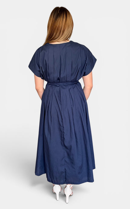 A woman viewed from the back wearing a navy blue midi dress with short sleeves, a gathered waist, and white high heels against a light gray studio background.