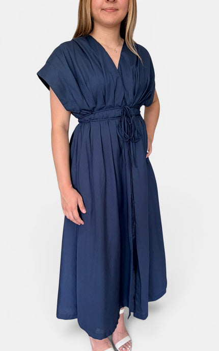 A woman wearing a navy blue midi dress with short flutter sleeves, a V-neckline, and a matching waist tie, shown against a plain light gray background.