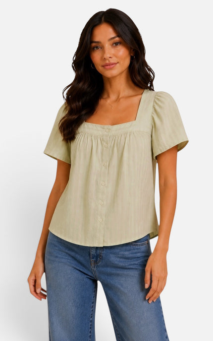 A woman modeling a light sage green short-sleeve blouse with a square neckline, puff sleeves, and a button-front smocked bodice, paired with blue jeans against a plain light gray studio background.