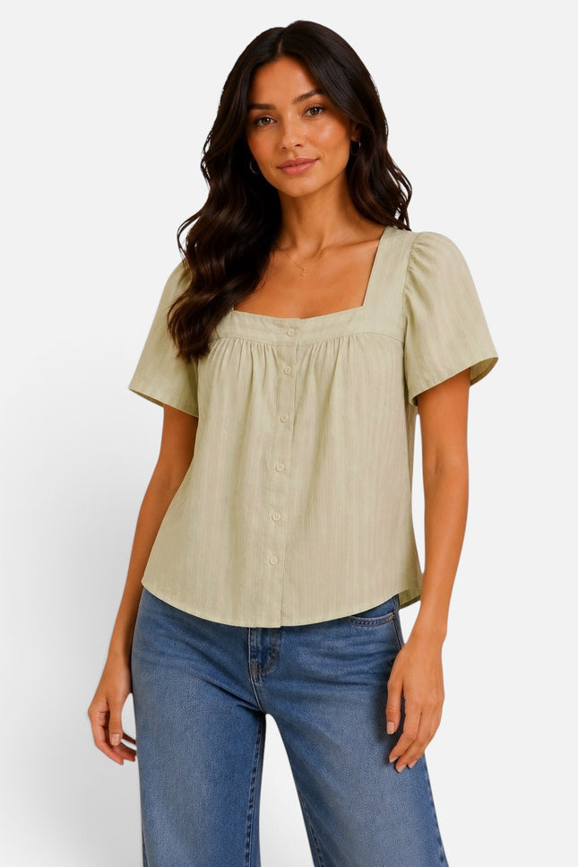 A woman modeling a light sage green short-sleeve blouse with a square neckline, puff sleeves, and a button-front smocked bodice, paired with blue jeans against a plain light gray studio background.