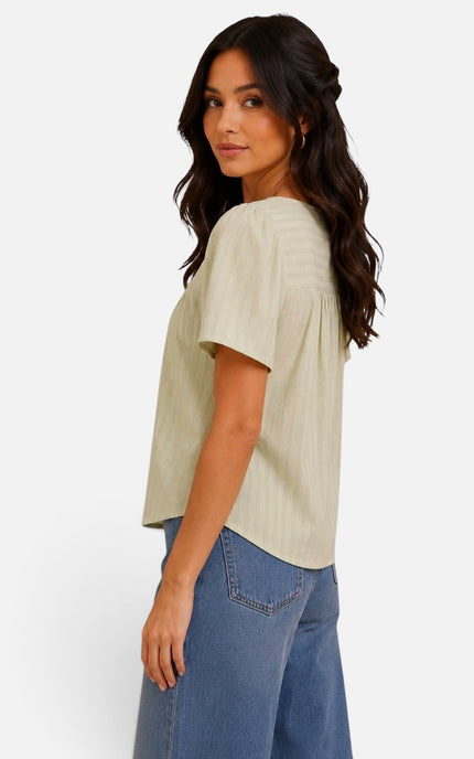 A woman wearing a light beige short-sleeve top with vertical pleats and blue jeans, shown from a side-back angle against a plain white background.
