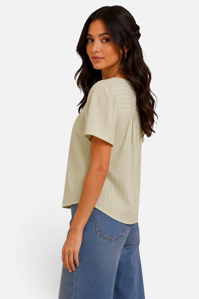 A woman wearing a light beige short-sleeve top with vertical pleats and blue jeans, shown from a side-back angle against a plain white background.