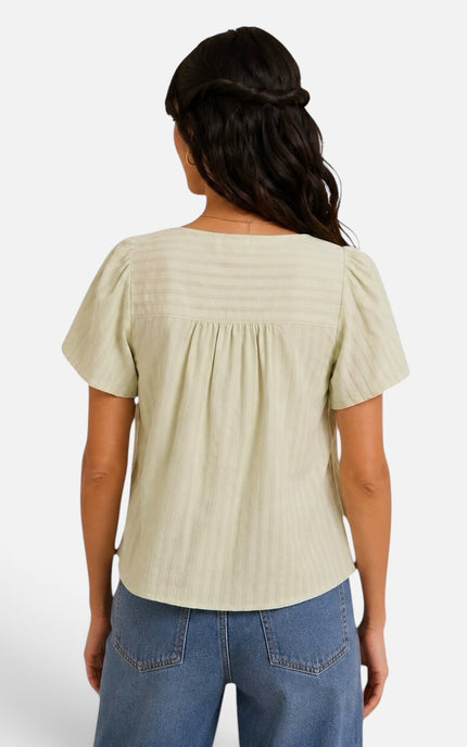 A woman viewed from the back wearing a short-sleeve, smocked-back top in a light sage green or pale yellow-green, paired with blue jeans, against a plain light gray studio background.