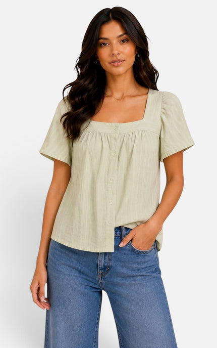A woman modeling a light sage green short-sleeve blouse with a square neckline and button-front detail, paired with blue jeans against a plain light gray studio background.