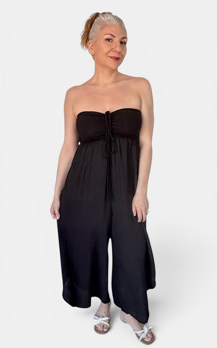 A woman modeling a black strapless wide-leg jumpsuit with a gathered sweetheart neckline and front tie detail, paired with white heeled sandals, against a light gray studio background.