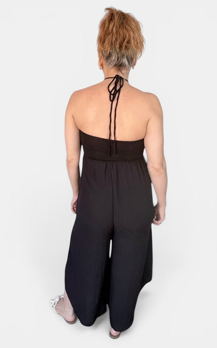 A woman viewed from the back wearing a black halter-style wide-leg jumpsuit with tie-back straps, standing against a light gray studio background.