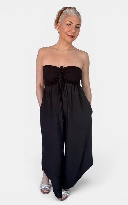 A woman modeling a strapless black jumpsuit with a gathered sweetheart-style bodice and wide legs, photographed against a plain light gray studio background.