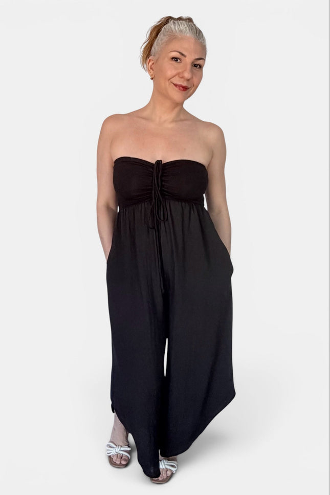 A woman modeling a strapless black jumpsuit with a gathered sweetheart-style bodice and wide legs, photographed against a plain light gray studio background.