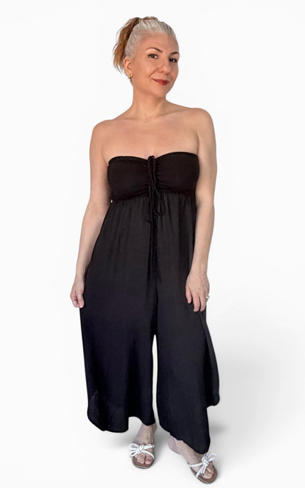 A woman modeling a black strapless jumpsuit with a ruched bodice and wide-leg pants, paired with white sandals, against a plain white studio background.