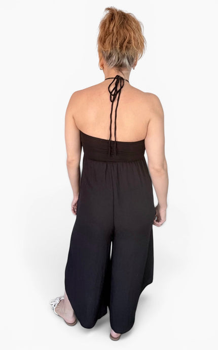 A woman viewed from behind wearing a black halter-neck jumpsuit with wide legs and tie-back straps, standing against a plain white studio background.