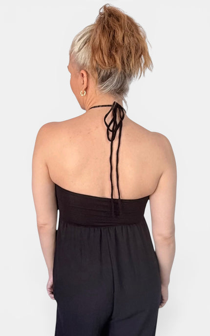 Rear view of a woman with blonde hair in a high ponytail wearing a black strapless halter jumpsuit with tie-back straps, set against a plain light gray background.