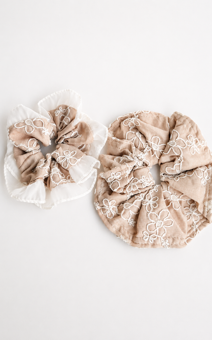 Two fabric scrunchies on a plain white background. One is beige with white floral embroidery and a white ruffled trim, and the other is a solid beige fabric with a subtle tonal floral pattern.