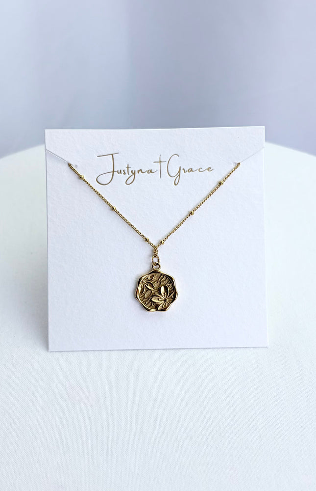An etched floral cameo coin necklace arranged on a clean boutique display card, showcasing the full pendant face and the tarnish-resistant satellite chain.