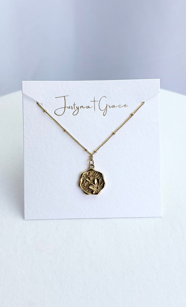 An etched floral cameo coin necklace arranged on a clean boutique display card, showcasing the full pendant face and the tarnish-resistant satellite chain.