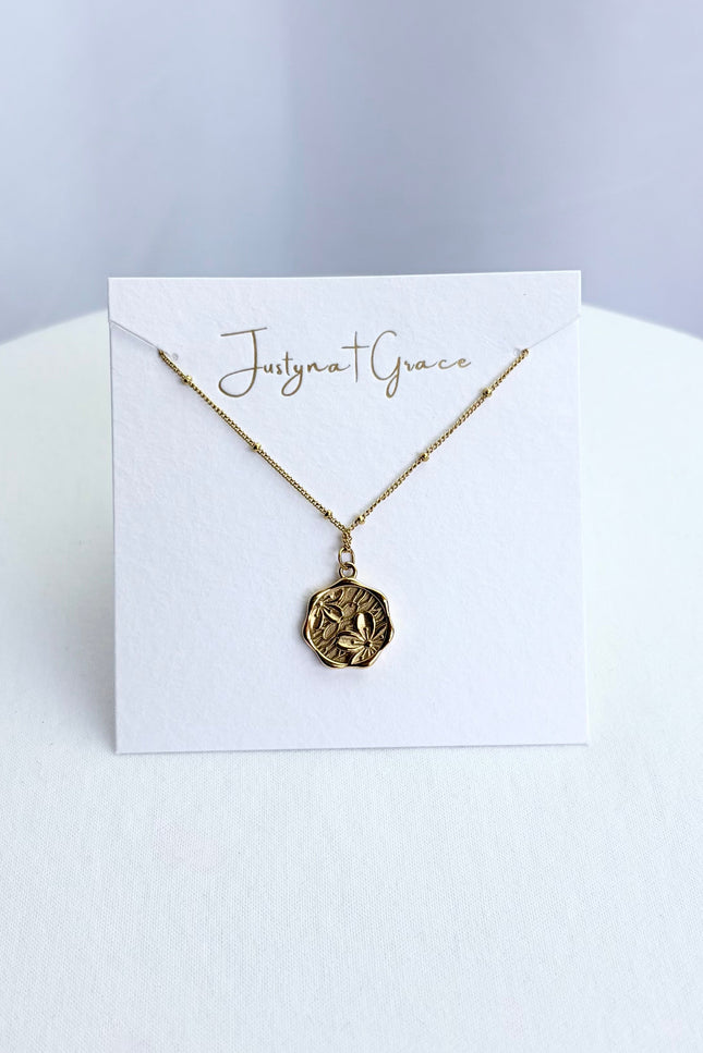 An etched floral cameo coin necklace arranged on a clean boutique display card, showcasing the full pendant face and the tarnish-resistant satellite chain.