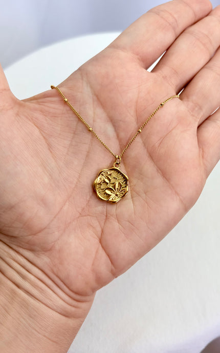A handheld close-up of a gold organic coin necklace resting in the palm of a hand. The shot highlights the intricately etched floral cameo design on the pendant face and the detailed texture of the delicate satellite chain.
