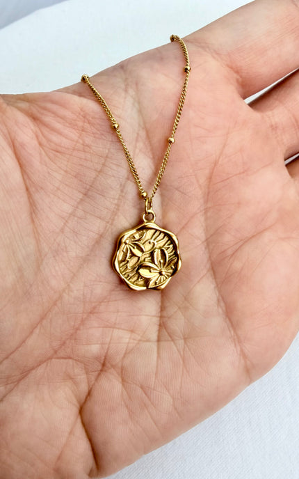 Close-up photo of the vintage-inspired floral cameo coin pendant held in an open palm, showcasing its textured, organic edges and the high-shine finish of the 18k gold PVD plating.