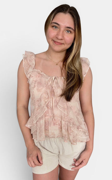A young woman modeling a light pink and beige ruffled tie-front top with flutter sleeves, paired with cream shorts, against a plain white background.