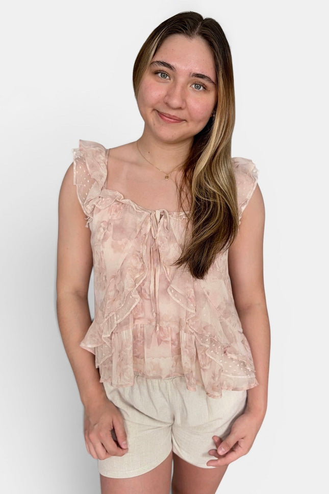 A young woman modeling a light pink and beige ruffled tie-front top with flutter sleeves, paired with cream shorts, against a plain white background.