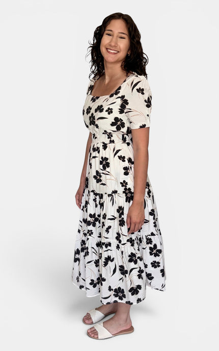 A woman modeling a short-sleeve midi dress with a black floral print on a white background, paired with white slide sandals, against a light gray studio backdrop.