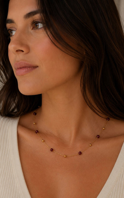 A close-up of a woman wearing a delicate gold necklace with small dark red beads, paired with a cream-colored ribbed top.