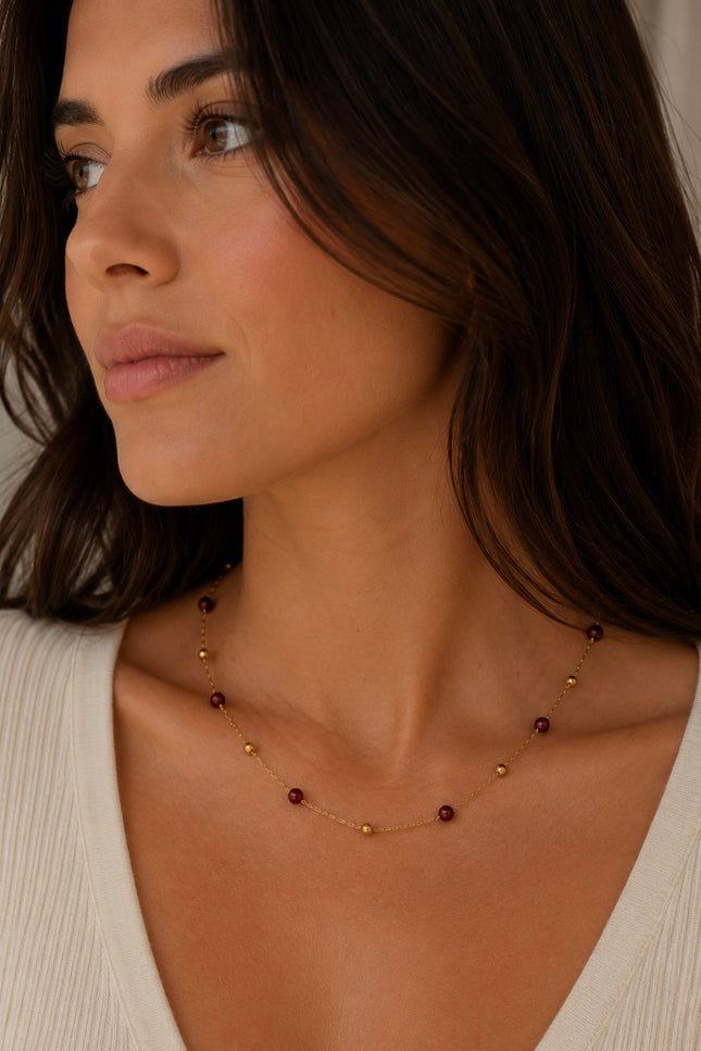 A close-up of a woman wearing a delicate gold necklace with small dark red beads, paired with a cream-colored ribbed top.