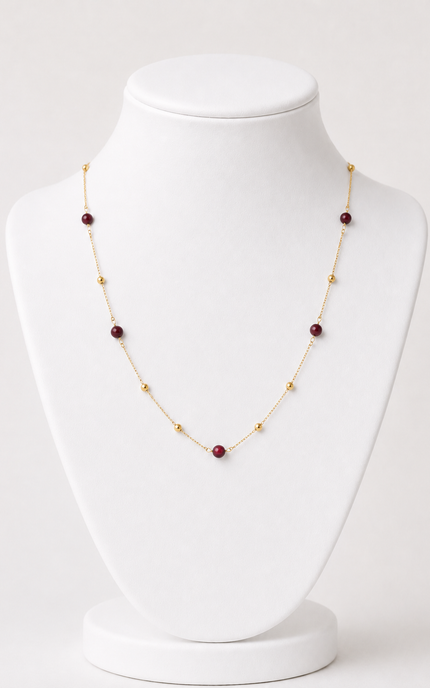 A delicate gold chain necklace with alternating small gold beads and round dark red gemstone beads, displayed on a white jewelry bust.