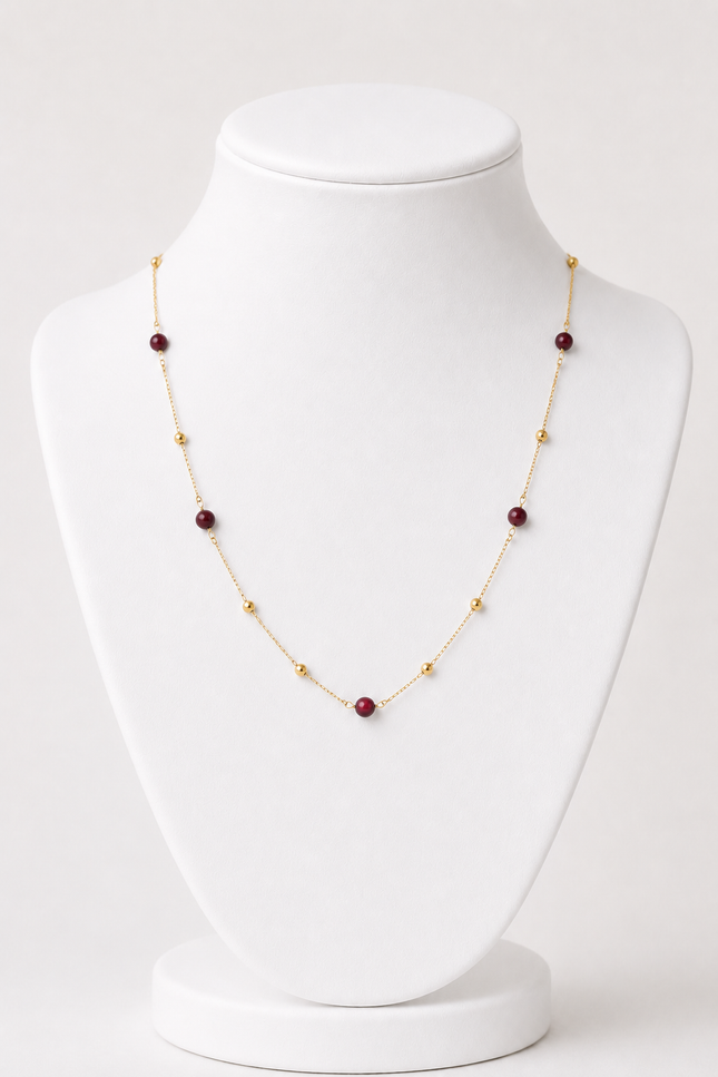 A delicate gold chain necklace with alternating small gold beads and round dark red gemstone beads, displayed on a white jewelry bust.