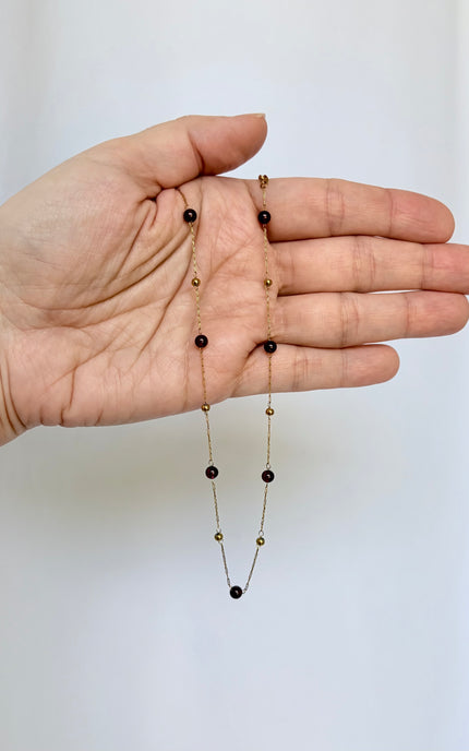 A delicate gold chain necklace with alternating small gold beads and dark round stones, draped across an open hand to show scale and detail.