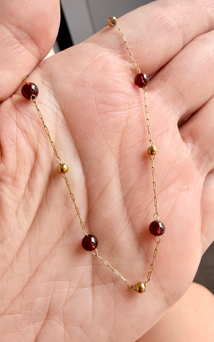 A delicate gold chain necklace with small dark red beads and a few polished gold spacer beads, held in a person's hand.