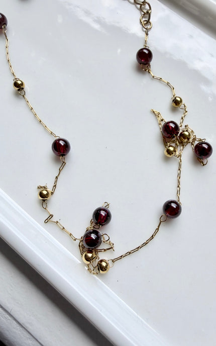 A delicate gold-tone chain necklace with round dark burgundy beads and small gold ball accents, displayed on a white surface.