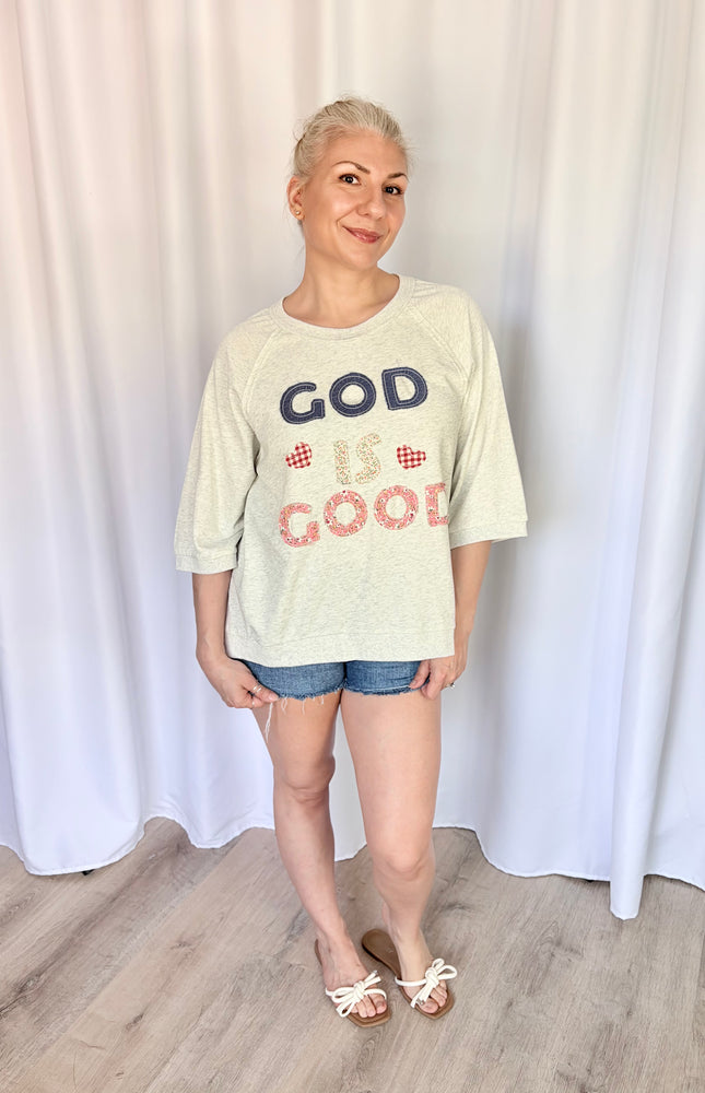 Front view of a model wearing a loose, casual French terry top featuring a vintage-inspired "God is Good" patchwork design across the chest and comfortable 3/4 length raglan sleeves.