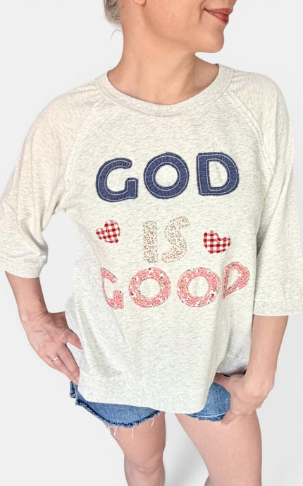 A woman wearing a heather cream 3/4 sleeve graphic t-shirt with the text 'GOD IS GOOD' in navy and red gingham hearts patchwork, paired with denim shorts.
