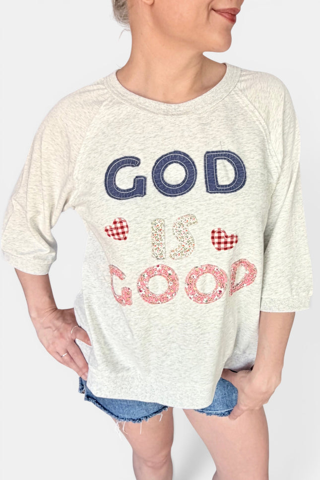 A woman wearing a heather cream 3/4 sleeve graphic t-shirt with the text 'GOD IS GOOD' in navy and red gingham hearts patchwork, paired with denim shorts.