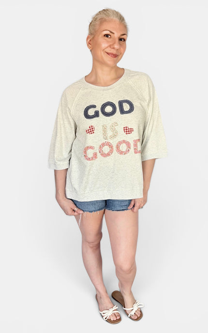 A woman modeling a cream-colored heathered t-shirt with 'GOD IS GOOD' patchwork on the front in navy and red patterned lettering, paired with denim shorts and white slide sandals, against a light gray studio background.