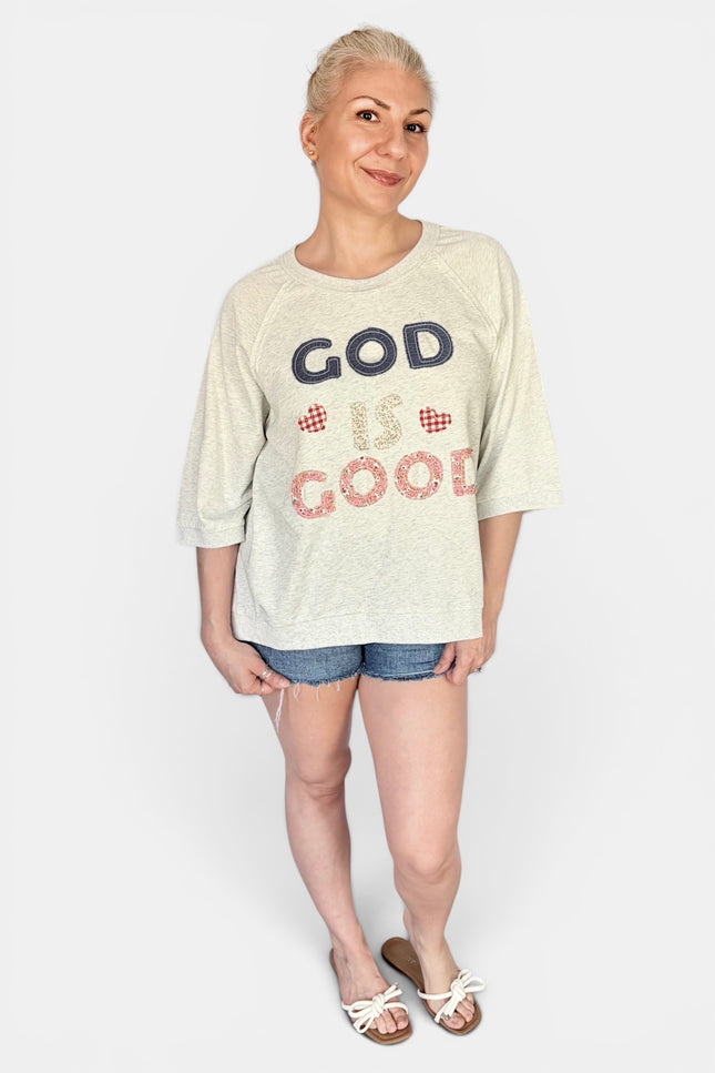 A woman modeling a cream-colored heathered t-shirt with 'GOD IS GOOD' patchwork on the front in navy and red patterned lettering, paired with denim shorts and white slide sandals, against a light gray studio background.