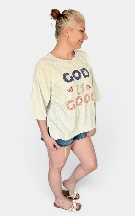 A woman modeling a cream-colored graphic t-shirt with 'GOD IS GOOD' patchwork lettering on the front in navy and pinkish-red, paired with denim shorts and sandals, against a light gray background.