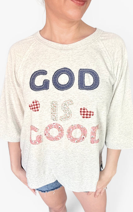 A woman wearing a light heather gray heather t-shirt with 'GOD IS GOOD' patchwork on the front in navy, pink, and red gingham patterns, paired with denim shorts.