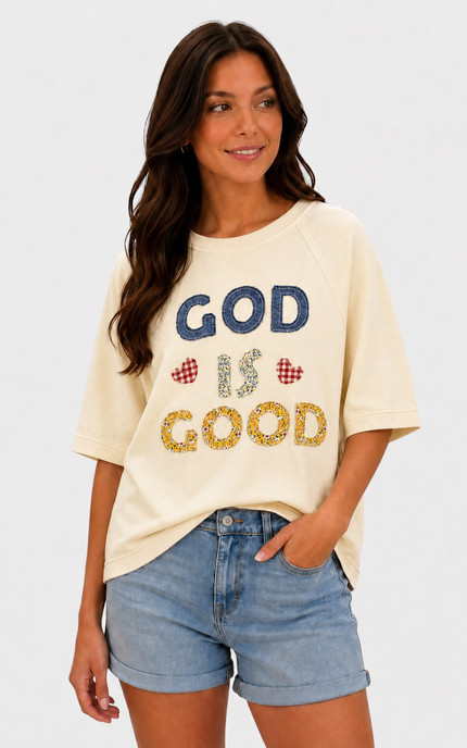 A woman wearing a cream-colored graphic t-shirt with 'GOD IS GOOD' written on the front in blue and yellow glitter-style lettering, paired with light-wash denim shorts, against a plain white background.