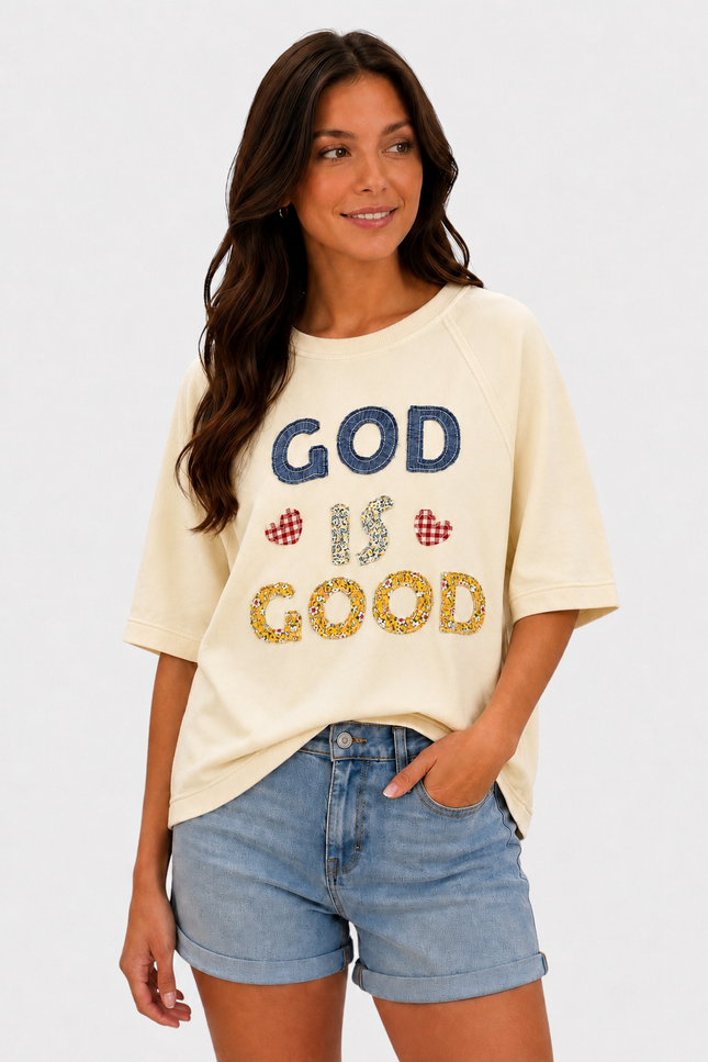 A woman wearing a cream-colored graphic t-shirt with 'GOD IS GOOD' written on the front in blue and yellow glitter-style lettering, paired with light-wash denim shorts, against a plain white background.