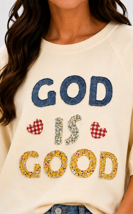 A close-up of a woman wearing a cream-colored sweatshirt with a decorative graphic that reads 'GOD IS GOOD' in blue denim, floral print, and red-and-white gingham hearts.